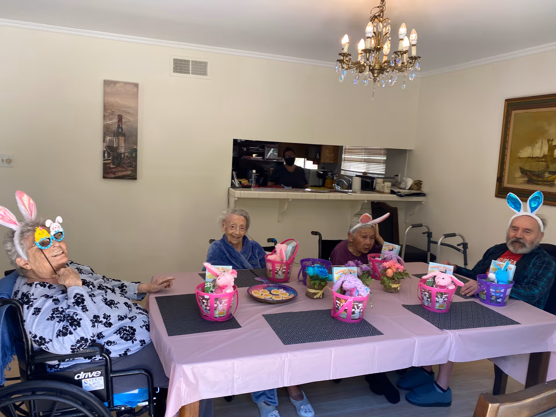Four elderly individuals sitting around a table decorated with pink tablecloth and Easter-themed baskets and flowers. Three of them are wearing bunny ears headbands and one is wearing Easter-themed glasses. In the background, a person wearing a mask is visible in the kitchen area behind a counter. The room has light-colored walls, a chandelier, and framed artwork.