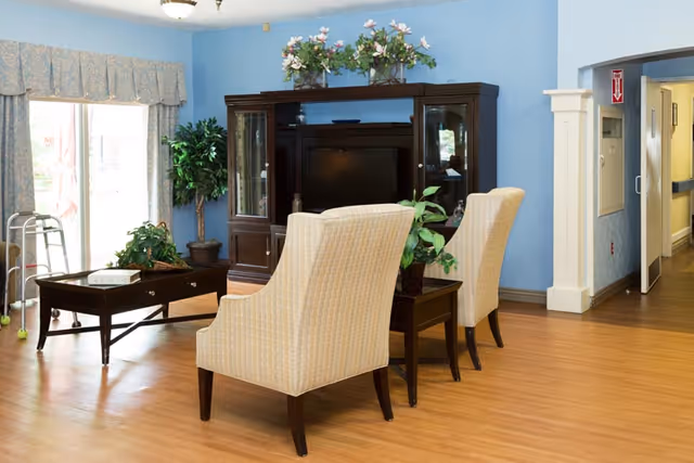 A cozy living room area in a senior care facility with two beige upholstered chairs facing a dark wooden entertainment center holding a television. The walls are painted light blue, and there are plants and floral decorations on top of the entertainment center. A coffee table and a walker are near a large window with patterned curtains, and a hallway is visible to the right.