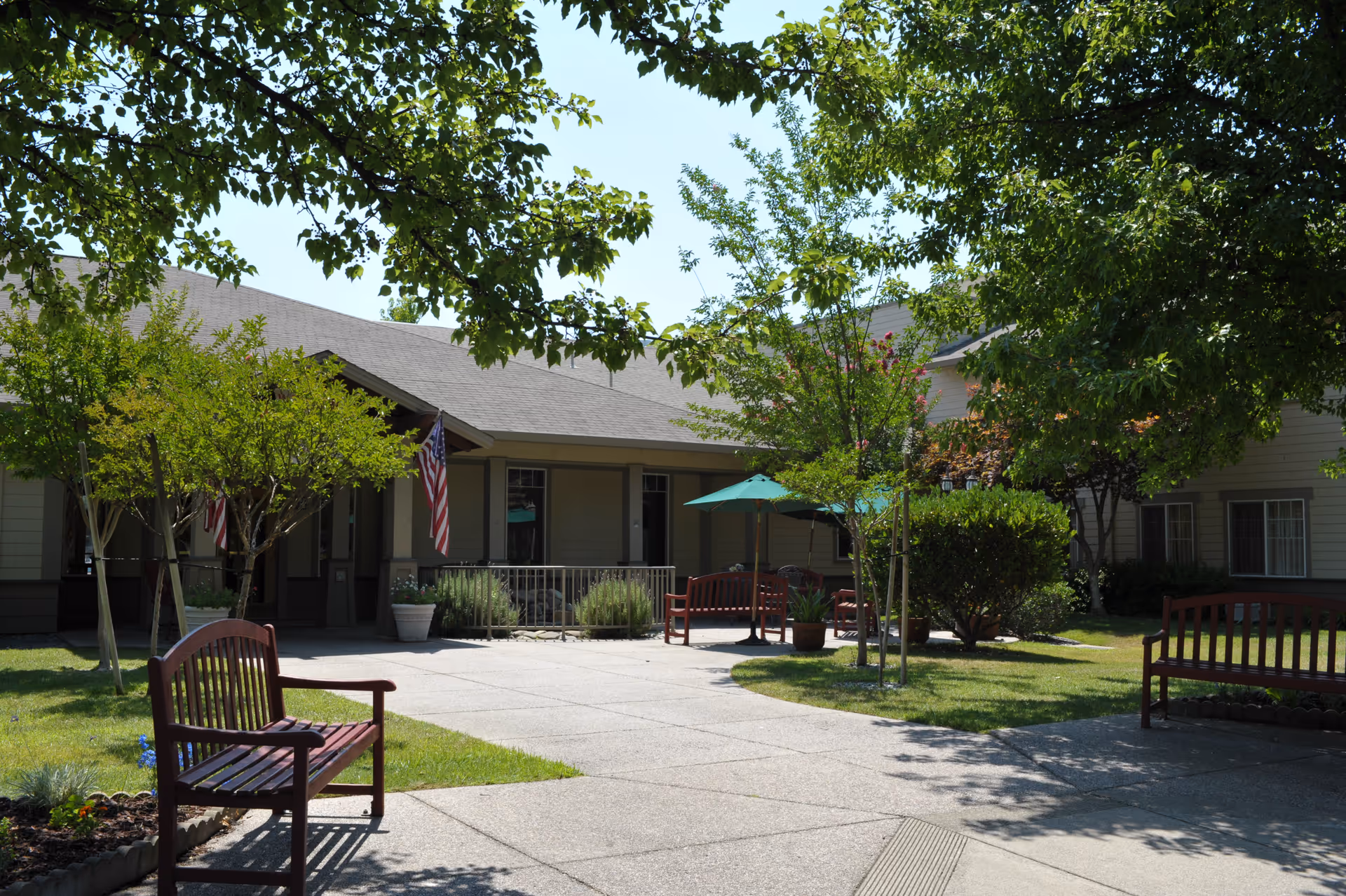 Outdoor courtyard area of Oakdale Heights Assisted Living featuring paved walkways, wooden benches, green trees, shrubs, and a building with American flags hanging near the entrance.