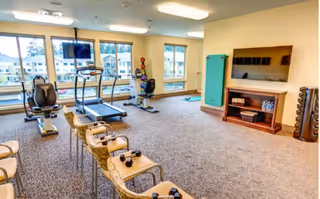A fitness room with exercise equipment including a treadmill, stationary bike, and recumbent bike. There are several chairs with small dumbbells placed on them, a wall-mounted TV, a shelf with water bottles, and large windows letting in natural light.