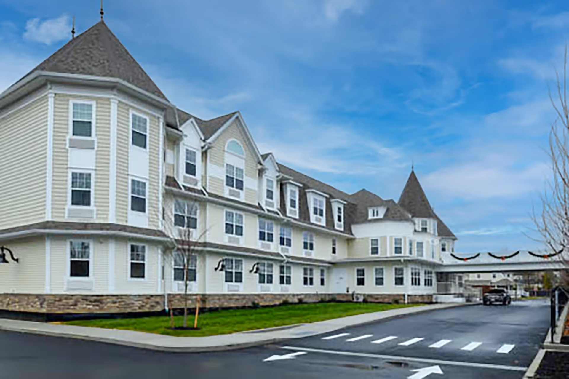 Exterior view of a large, multi-story senior living facility building with beige siding, multiple windows, and a steeply pitched roof with turrets. The building is surrounded by a paved driveway and a small grassy area with young trees under a partly cloudy blue sky.