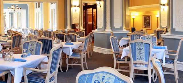 Elegant dining room with multiple tables covered in white tablecloths and blue napkins, surrounded by ornate chairs with blue and gold upholstery. The room features decorative columns, warm lighting, and framed artwork on the walls.