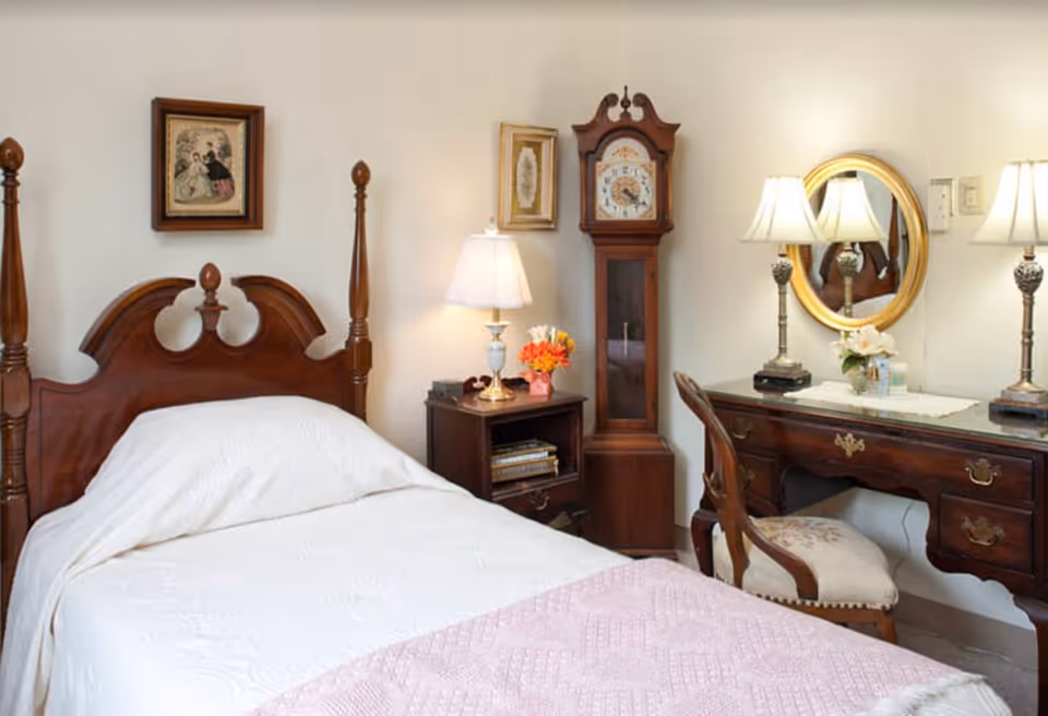 A cozy bedroom with a wooden four-poster bed covered in white bedding and a pink quilt at the foot. Next to the bed is a wooden nightstand with a lamp and a small flower arrangement. A tall grandfather clock stands beside the nightstand. On the right side, there is a wooden vanity desk with two lamps, a round mirror, and a cushioned chair with floral upholstery. The walls are decorated with framed artwork.