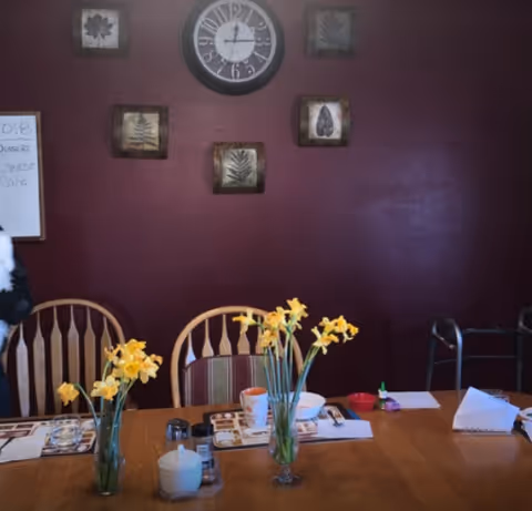 A dining room with a wooden table set with placemats, cups, and bowls. Two vases with yellow flowers are placed on the table. Behind the table are wooden chairs and a maroon wall decorated with a large clock and framed leaf artwork. A whiteboard with writing is partially visible on the left side, and a walker is seen on the right side.