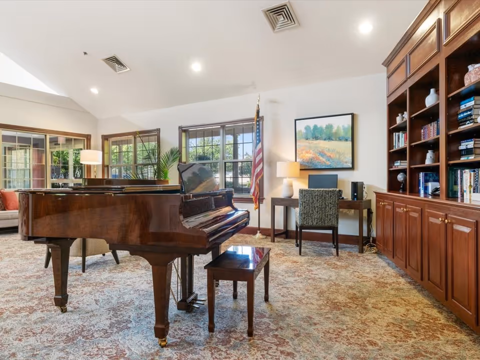 A bright common room featuring a grand piano, wooden bookshelves, a desk and chair, and seating by large windows.