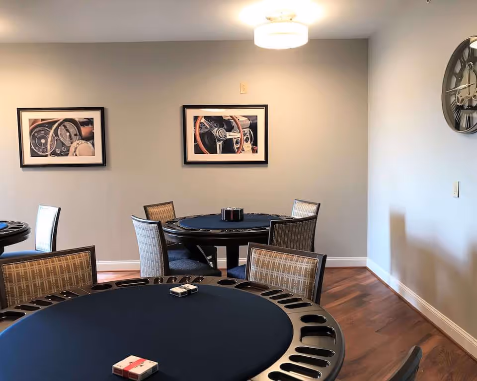 A room with round tables covered in dark blue felt, surrounded by chairs with patterned upholstery. On the tables are card decks and holders, suggesting a card or game room. The walls are light gray with two framed pictures of vintage car steering wheels and a large wall clock. The floor is wooden with a warm tone.