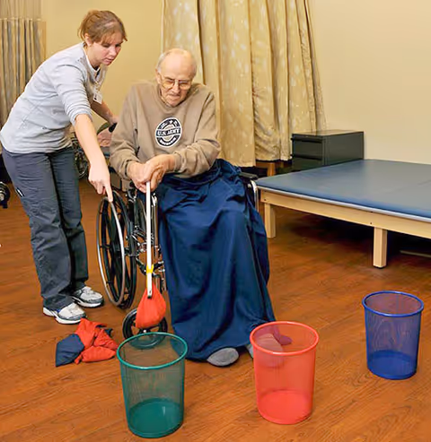 A caregiver assists an elderly man in a wheelchair as he plays a game tossing bean bags into colored bins in a room with wooden flooring, a padded table, and beige curtains.