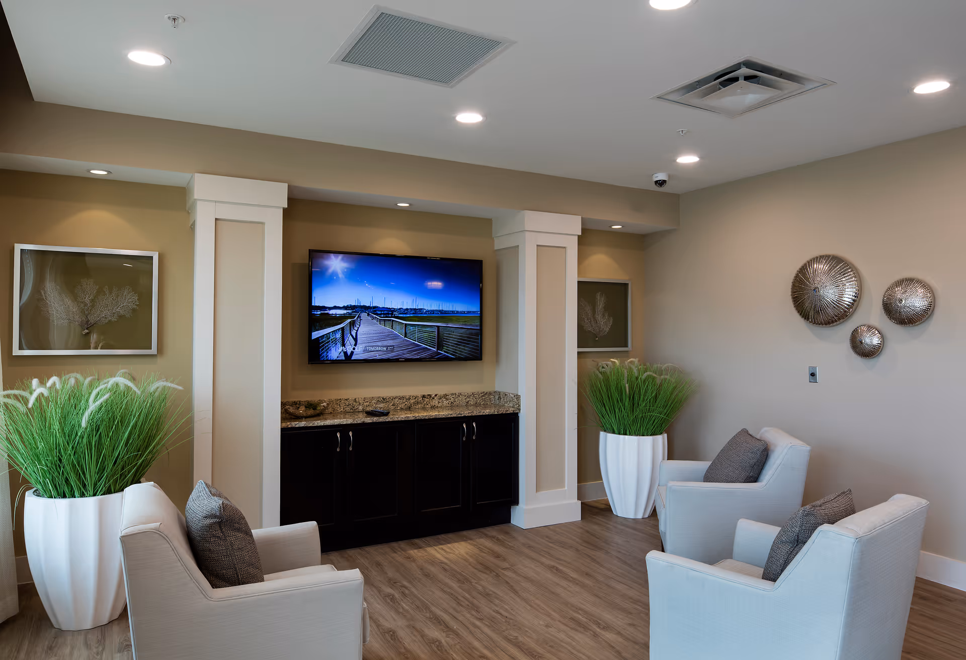 A cozy sitting area with four light-colored armchairs arranged around a wall-mounted flat-screen TV. The room features wood flooring, beige walls, decorative wall art, two large white planters with green plants, and recessed ceiling lights.