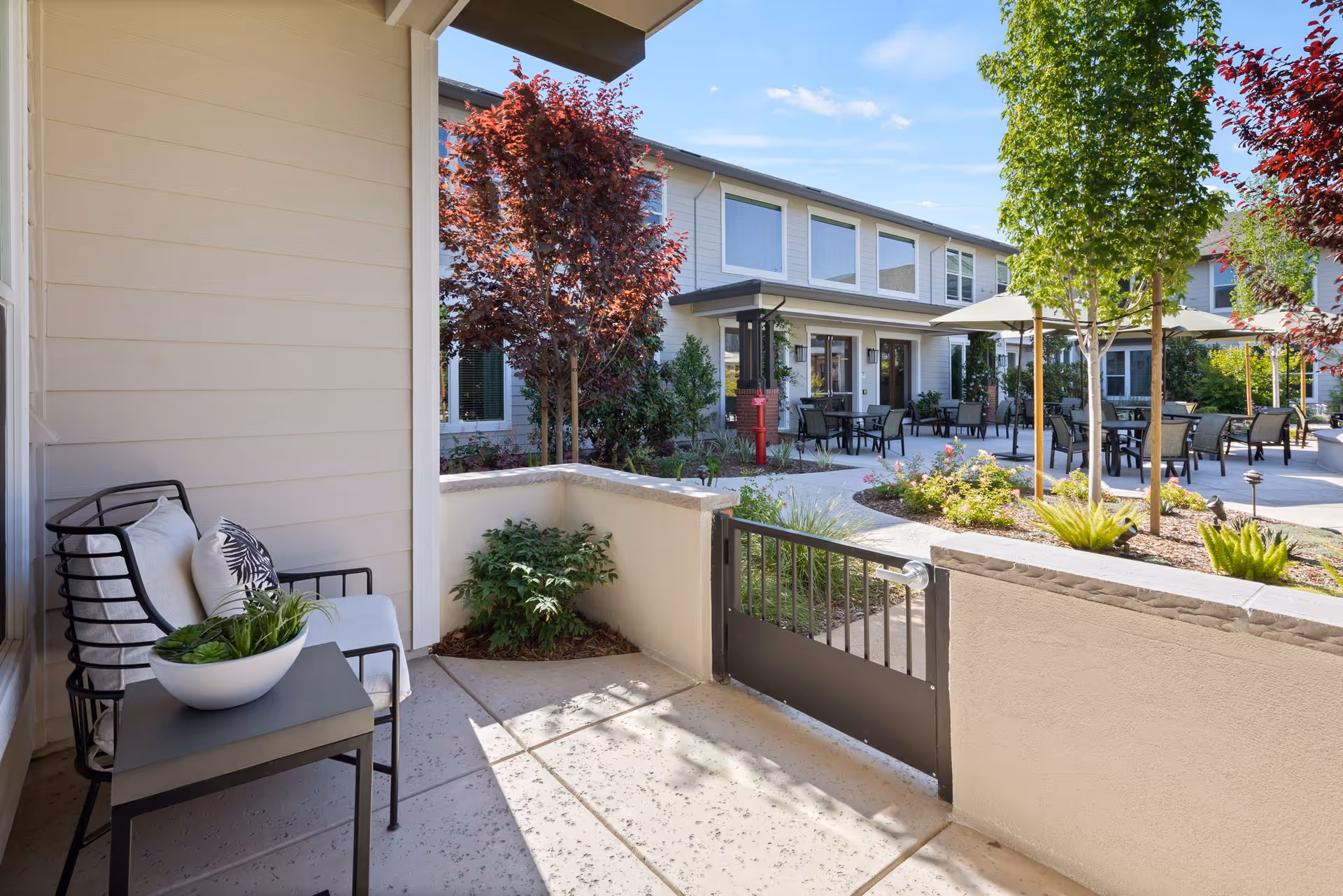 Small private patio with a chair, side table and potted plant overlooking a landscaped courtyard with outdoor dining tables and the building beyond.