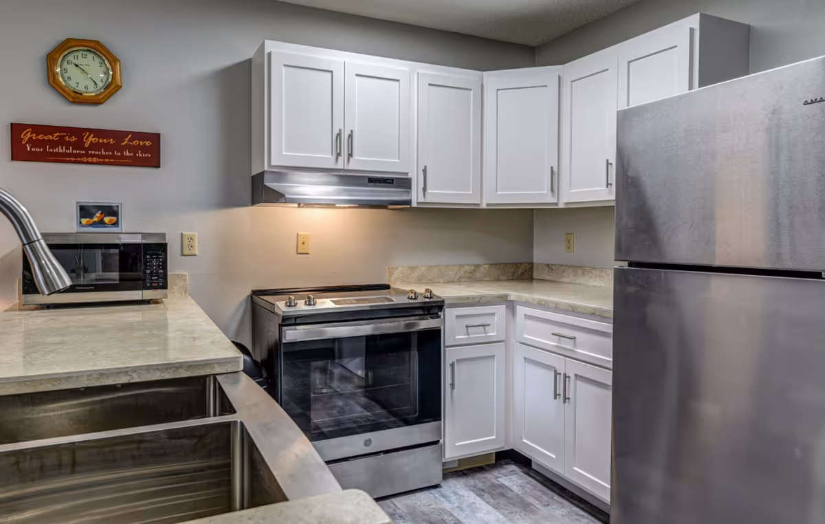 A modern kitchen with white cabinets, a stainless steel refrigerator, an electric stove with an overhead vent, a microwave on the counter, and a double sink. A wooden clock and a decorative sign with the text 'Great is Your Love Your faithfulness reaches to the skies' are mounted on the wall.