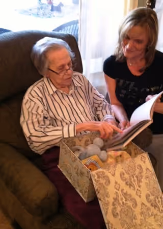 An elderly woman sitting in a cushioned armchair looking at a book with a younger woman beside her. The elderly woman is wearing glasses and a striped shirt, and there is a decorative box with soft toys on her lap. They appear to be engaged in a shared activity in a cozy indoor setting.