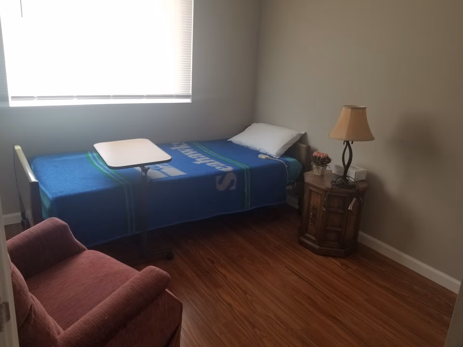 A small bedroom with a single bed covered with a blue blanket, a white pillow, and an overbed table. Next to the bed is a wooden nightstand with a lamp, a small flower pot, and a box of tissues. There is a red upholstered armchair in the foreground and a window with blinds letting in natural light.