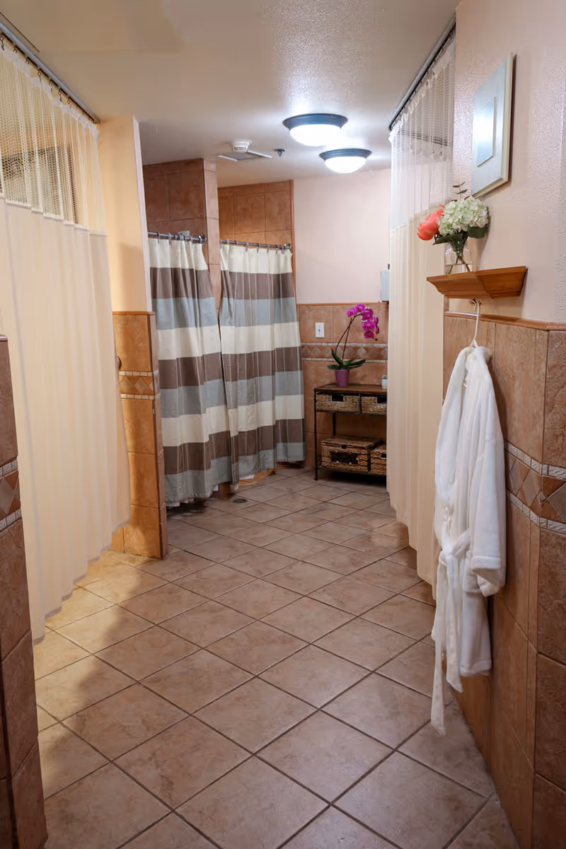 Tiled communal shower area with striped shower curtains, a hanging white robe, and a shelving unit with flowers and baskets.