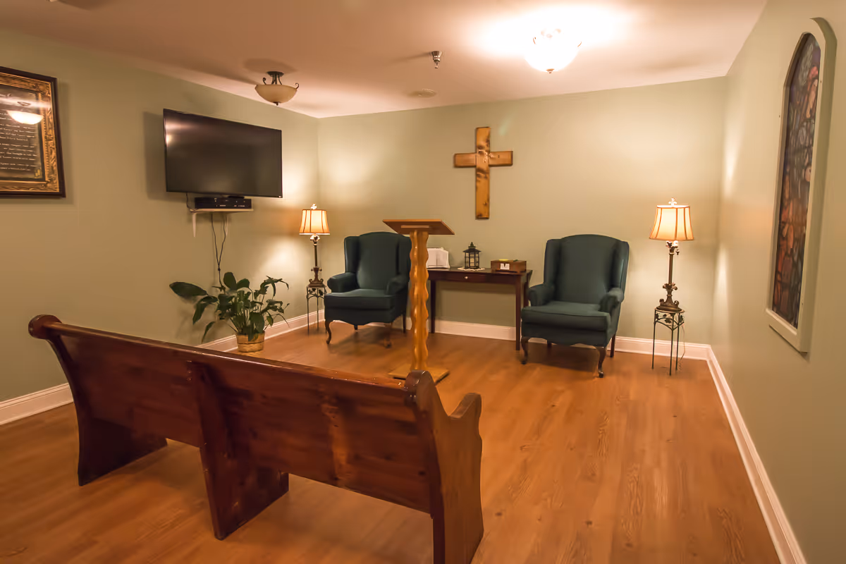 A small chapel room with wooden pews, two green armchairs, a wooden lectern, and a table with religious items. The walls are light green with a wooden cross mounted on the wall, a flat-screen TV, two lamps, a framed picture, and a potted plant on the floor.