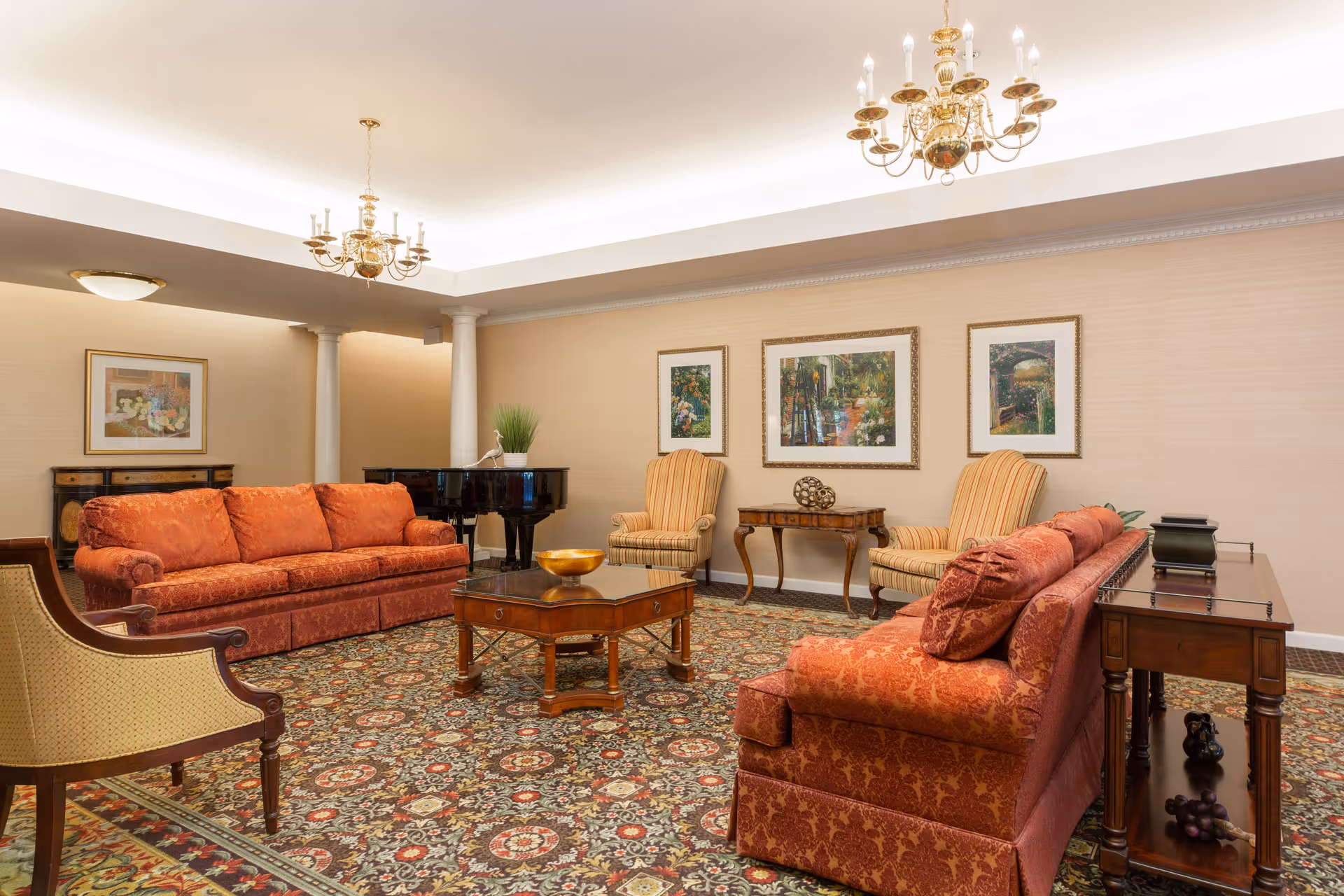 A spacious living room with two red patterned sofas, two striped armchairs, a wooden coffee table, and a side table. The room features a patterned carpet, framed artwork on the walls, two chandeliers, and a black grand piano in the corner.