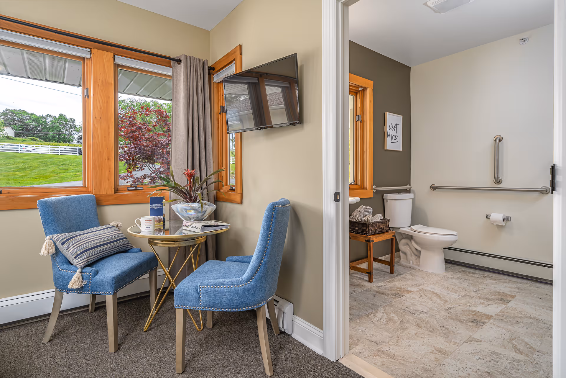 A cozy sitting area with two blue upholstered chairs and a small round table with a plant, book, and coffee mug, next to large windows with a view of green grass and trees. Adjacent to the sitting area is a bathroom with a toilet, grab bars, a small wooden table with a basket, and a framed picture on the wall.