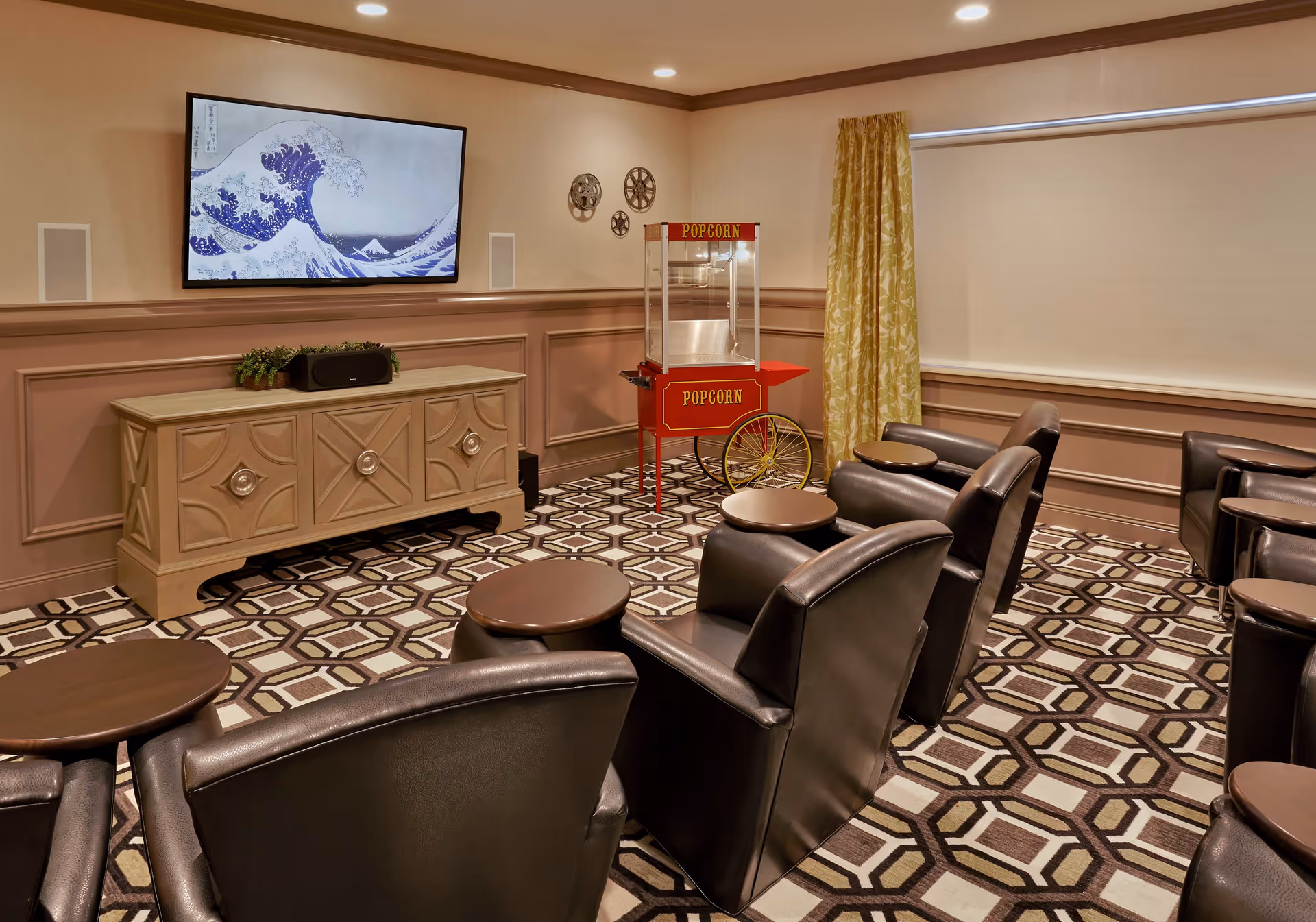 Small theater-style common room with leather recliner seats facing a wall-mounted TV, a popcorn machine, and a decorative cabinet.