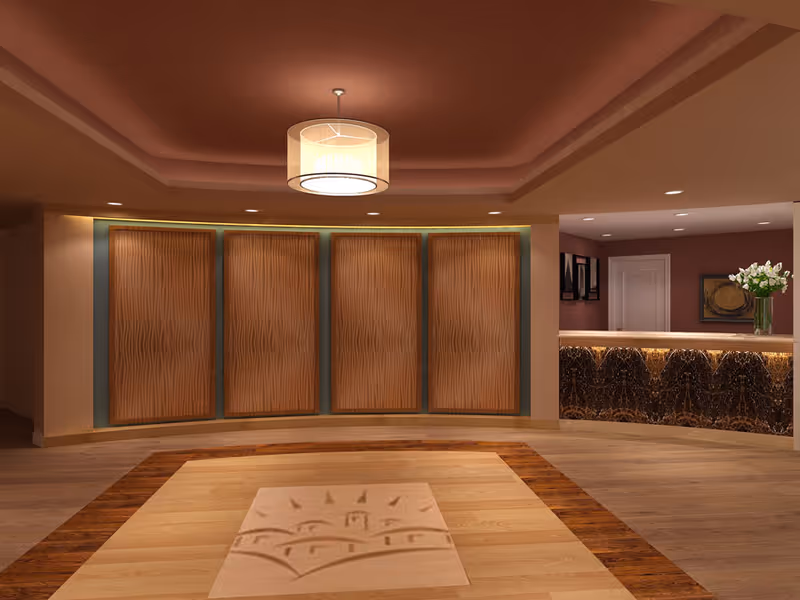Interior view of a senior living facility lobby area with a modern ceiling light fixture, four wooden decorative panels on the wall, a polished wooden floor with a patterned inlay, and a reception desk with a dark marble front and a vase of white flowers on top.