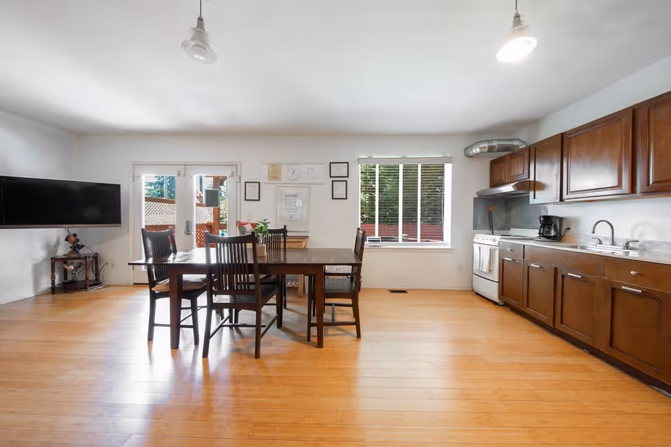 A bright and spacious room featuring a wooden dining table with six chairs in the center. To the right, there is a kitchen area with wooden cabinets, a stove, a coffee maker, and a sink. On the left wall, a flat-screen TV is mounted above a small table. The room has light-colored wooden flooring, a window with blinds, and a glass door leading outside.
