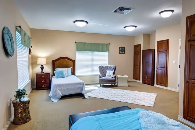 A spacious bedroom with two beds, one single bed with a wooden headboard and white bedding, and another bed partially visible with a blue blanket. The room has beige walls, carpeted floor, two windows with green valances, a wooden nightstand with a lamp, a comfortable armchair with a pillow, a small side table, and built-in wooden cabinets along one wall.