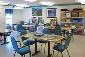 Bright communal activity room with several tables and blue chairs, a jigsaw puzzle on one table, shelving with storage bins, and framed pictures on the walls.