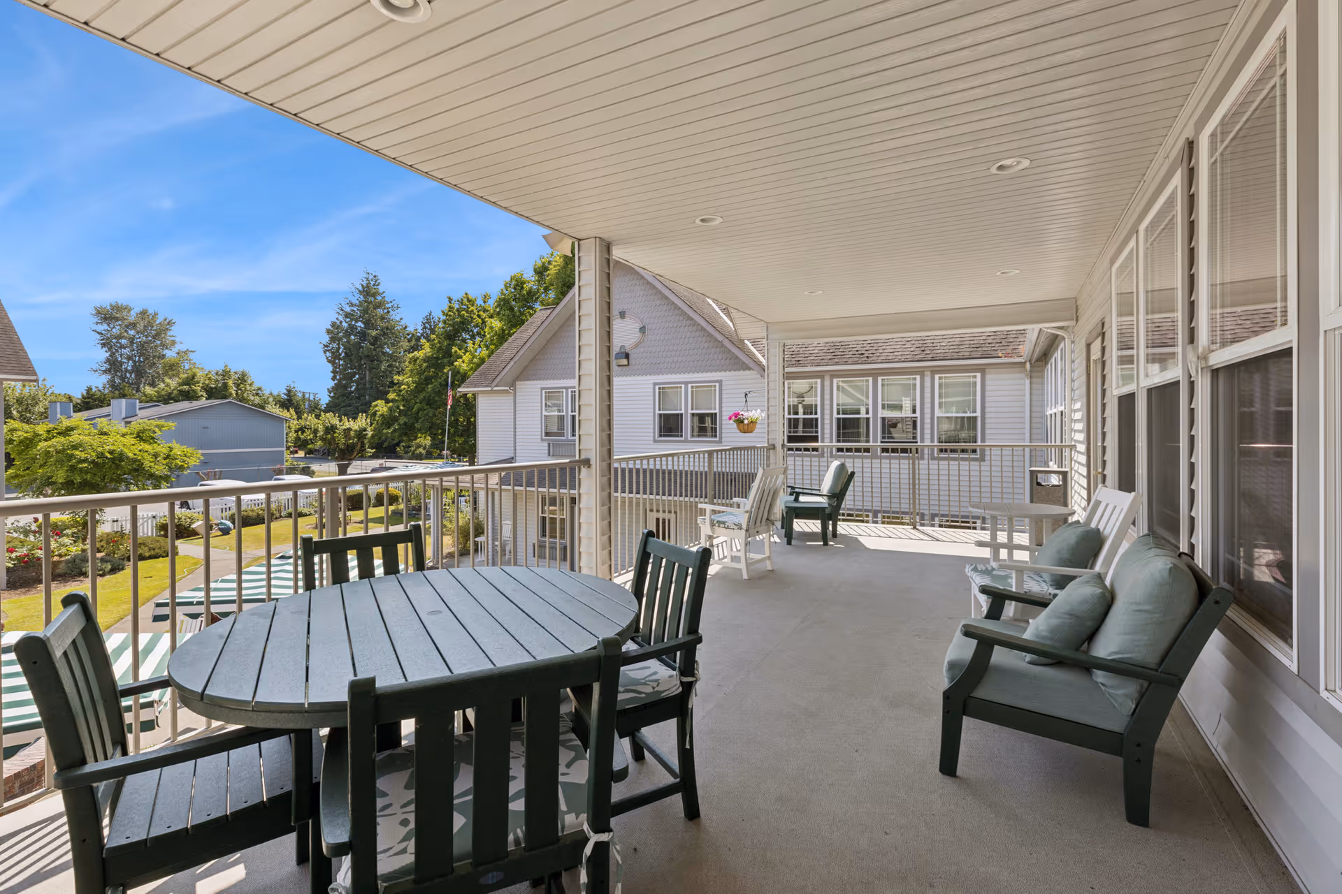Covered outdoor balcony area with a round table and four chairs, two cushioned armchairs, and additional seating. The balcony overlooks a garden area with trees and buildings under a clear blue sky.