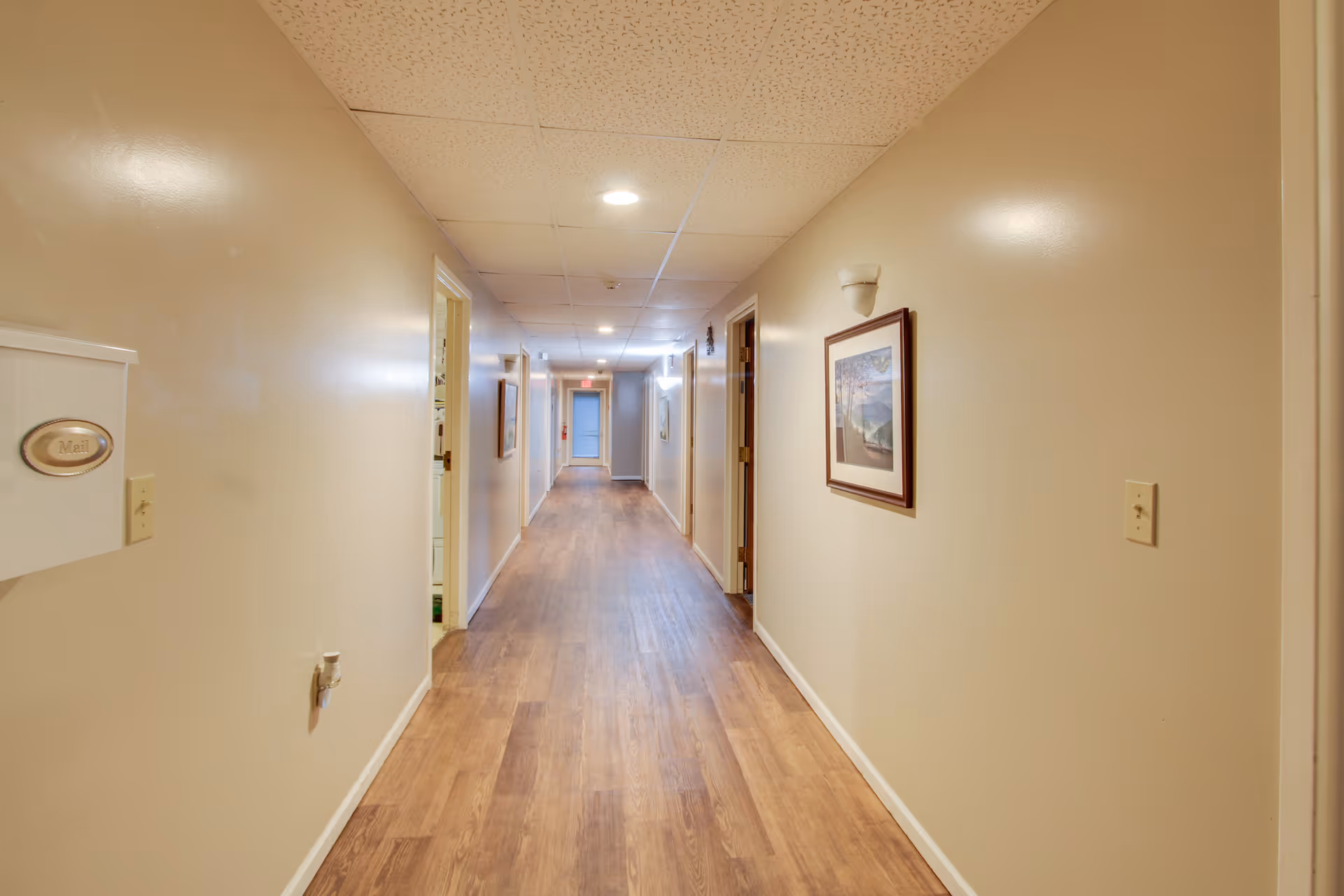 A long, narrow hallway with beige walls and wood flooring. Several doors line both sides of the hallway, and framed pictures hang on the walls. The ceiling has recessed lighting and a drop ceiling with white tiles. A white mailbox labeled 'Mail' is mounted on the left wall near the foreground.