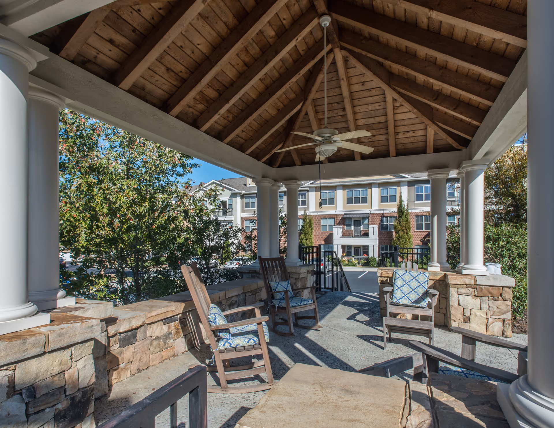 Covered outdoor seating area with wooden rocking chairs and stone walls under a wooden roof with a ceiling fan, overlooking a parking lot and a multi-story residential building in the background.