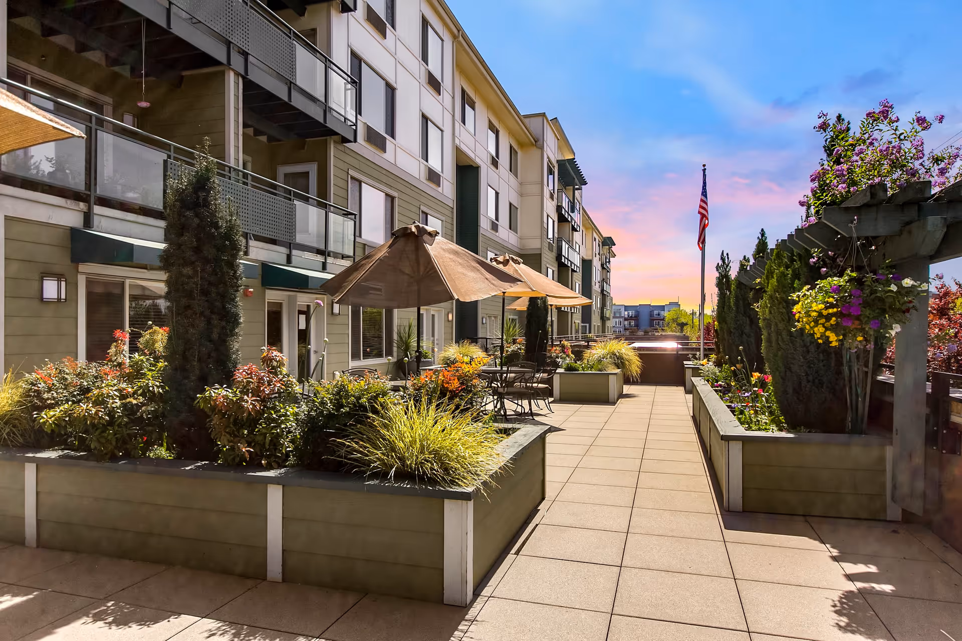 Outdoor patio area at Merrill Gardens at Renton Centre featuring raised garden beds with various plants and flowers, patio tables with umbrellas, and an American flag against a colorful sunset sky.