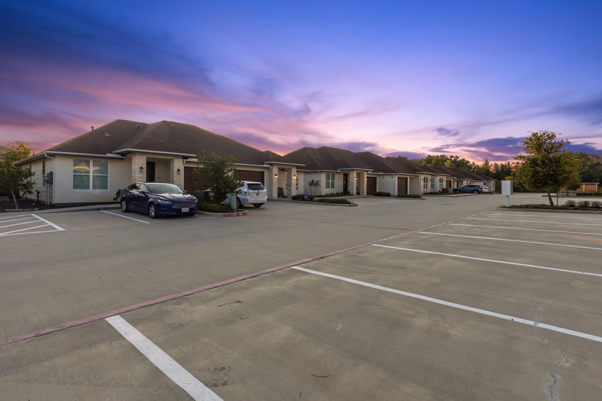 Row of single-story senior living units with parked cars and a large empty parking lot under a purple sunset sky.
