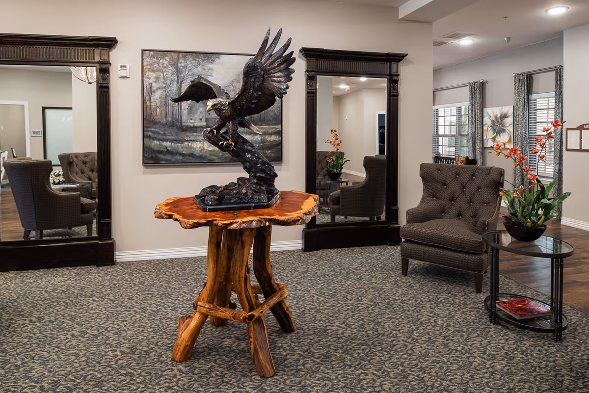 A cozy senior living facility common area with a rustic wooden table featuring a large eagle sculpture. The room has patterned carpet, two large dark-framed mirrors on the wall, a comfortable upholstered armchair, a glass side table with a floral arrangement, and windows with curtains letting in natural light.