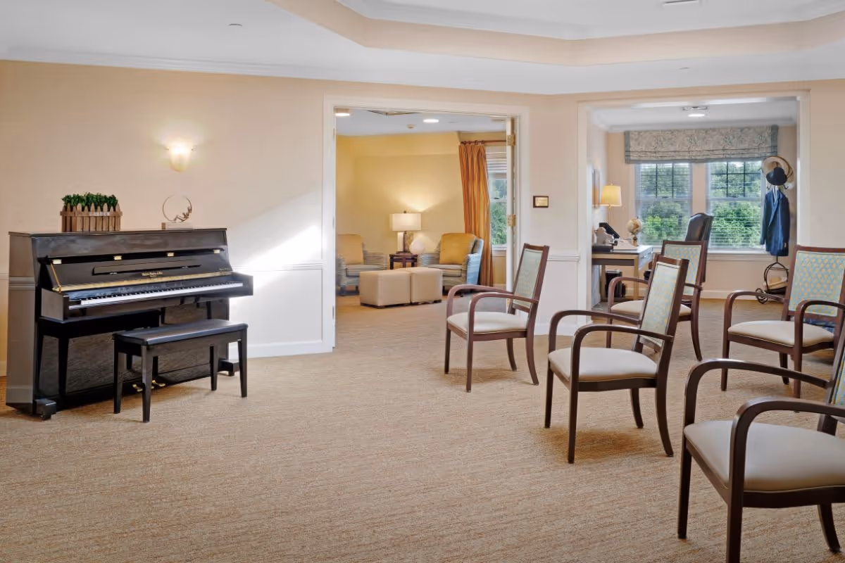 A bright and spacious common area in a senior living facility featuring a black upright piano with a bench on the left, several wooden chairs with cushioned seats arranged on the right, and an open doorway leading to a cozy sitting room with armchairs, ottomans, and a lamp. Another room with a desk, chair, coat rack, and large windows with a view of greenery is visible in the background.