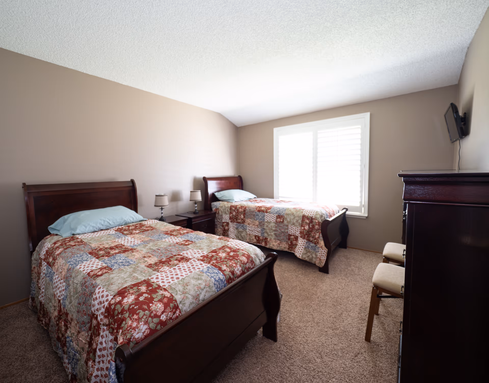 A bedroom with two twin beds featuring wooden frames and patchwork quilts in red, blue, and beige tones. Between the beds is a small wooden nightstand with two matching lamps. The room has beige walls, a carpeted floor, a window with white shutters, a wall-mounted TV, and a wooden dresser with two chairs beside it.