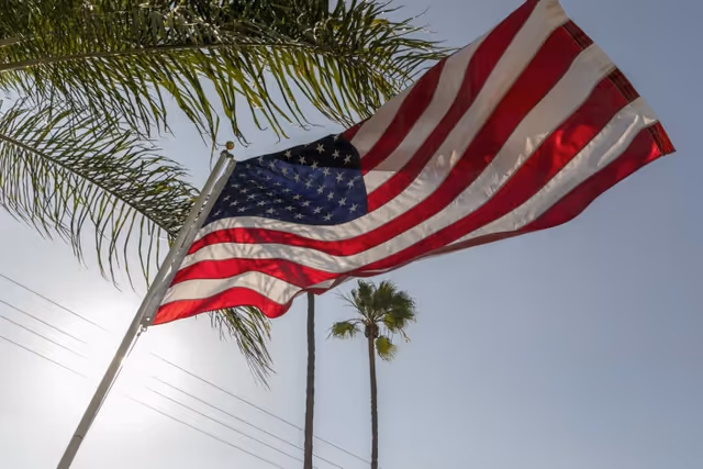 An American flag waves on a pole against a clear sky with palm trees in the background.