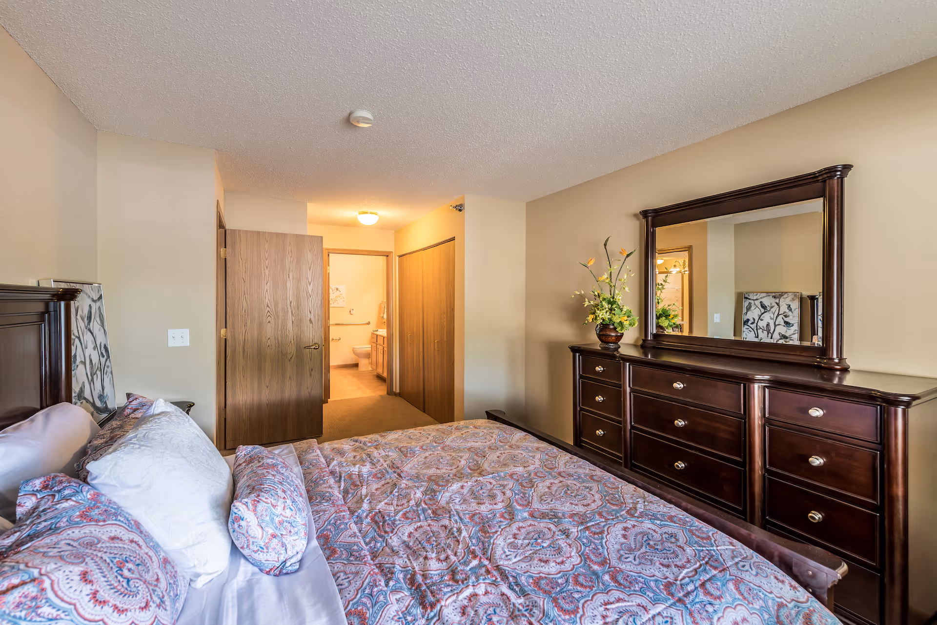 A bedroom with a bed covered in a patterned bedspread and multiple pillows. There is a wooden dresser with a large mirror and a vase with flowers on top. An open door reveals a bathroom with a toilet and sink visible. The walls are painted beige and the room has a carpeted floor.
