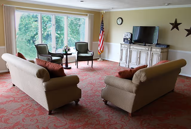 A bright communal living room with two sofas facing a TV, two chairs by a large window, and an American flag in the corner.