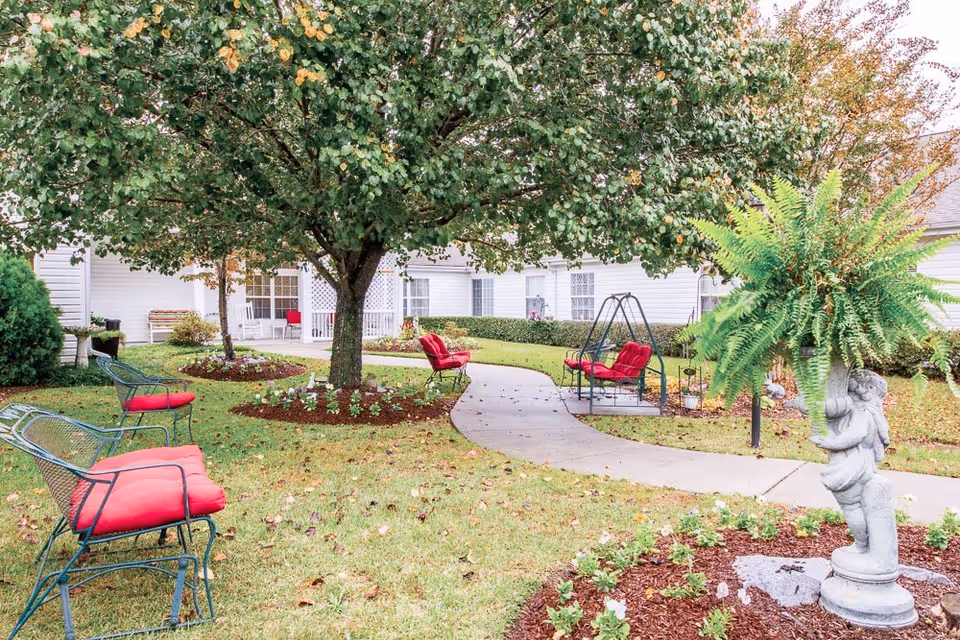 Landscaped courtyard with a large tree, red-cushioned benches, a swing and a cherub planter beside a curved concrete walkway in front of a white building.