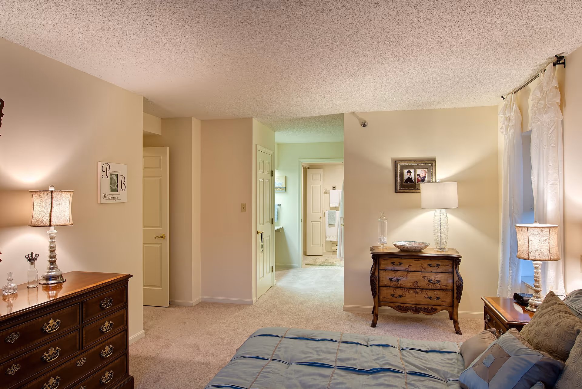 A cozy bedroom in a senior living facility with a bed covered in a blue comforter and multiple pillows. The room features two wooden dressers with lamps on top, a framed picture on the wall, and light-colored walls and carpet. A doorway leads to a bathroom area with towels visible.