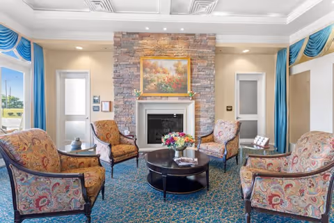 A cozy sitting area with four upholstered armchairs arranged around a round black coffee table with a flower arrangement on top. The room features a stone fireplace with a framed floral painting above it, two doors with frosted glass panels on either side of the fireplace, and large windows with blue curtains letting in natural light.