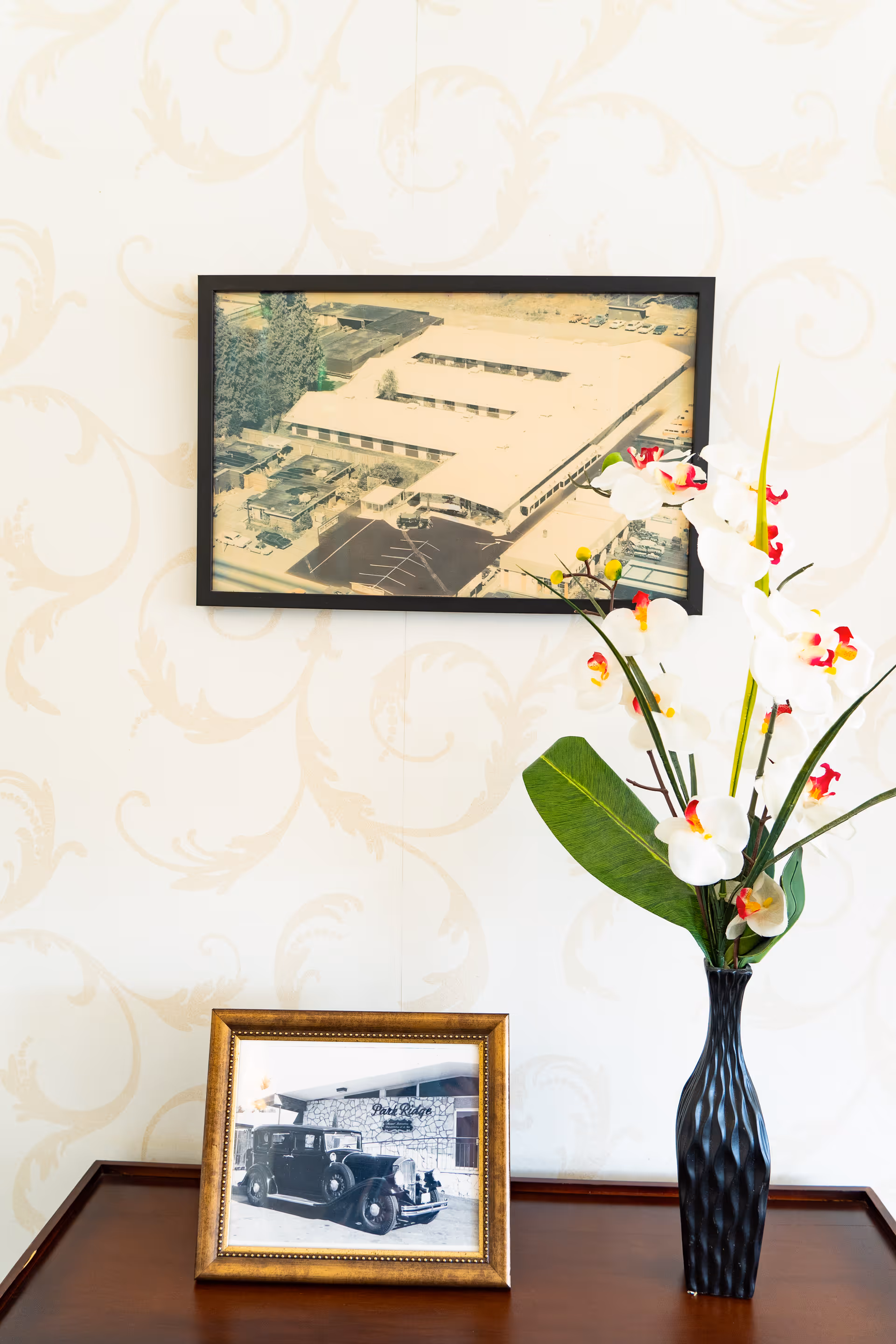Framed aerial photo and a vintage car picture on a wooden table next to a vase of white orchids against patterned wallpaper.