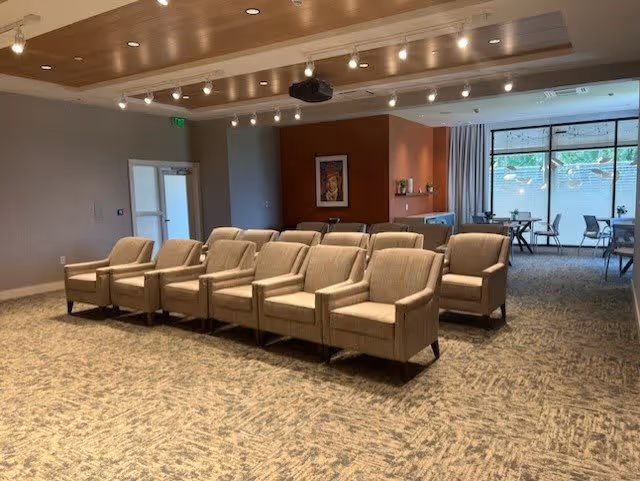 A spacious common room with two rows of upholstered chairs facing a projector screen area, recessed ceiling lights, and tables by large windows.