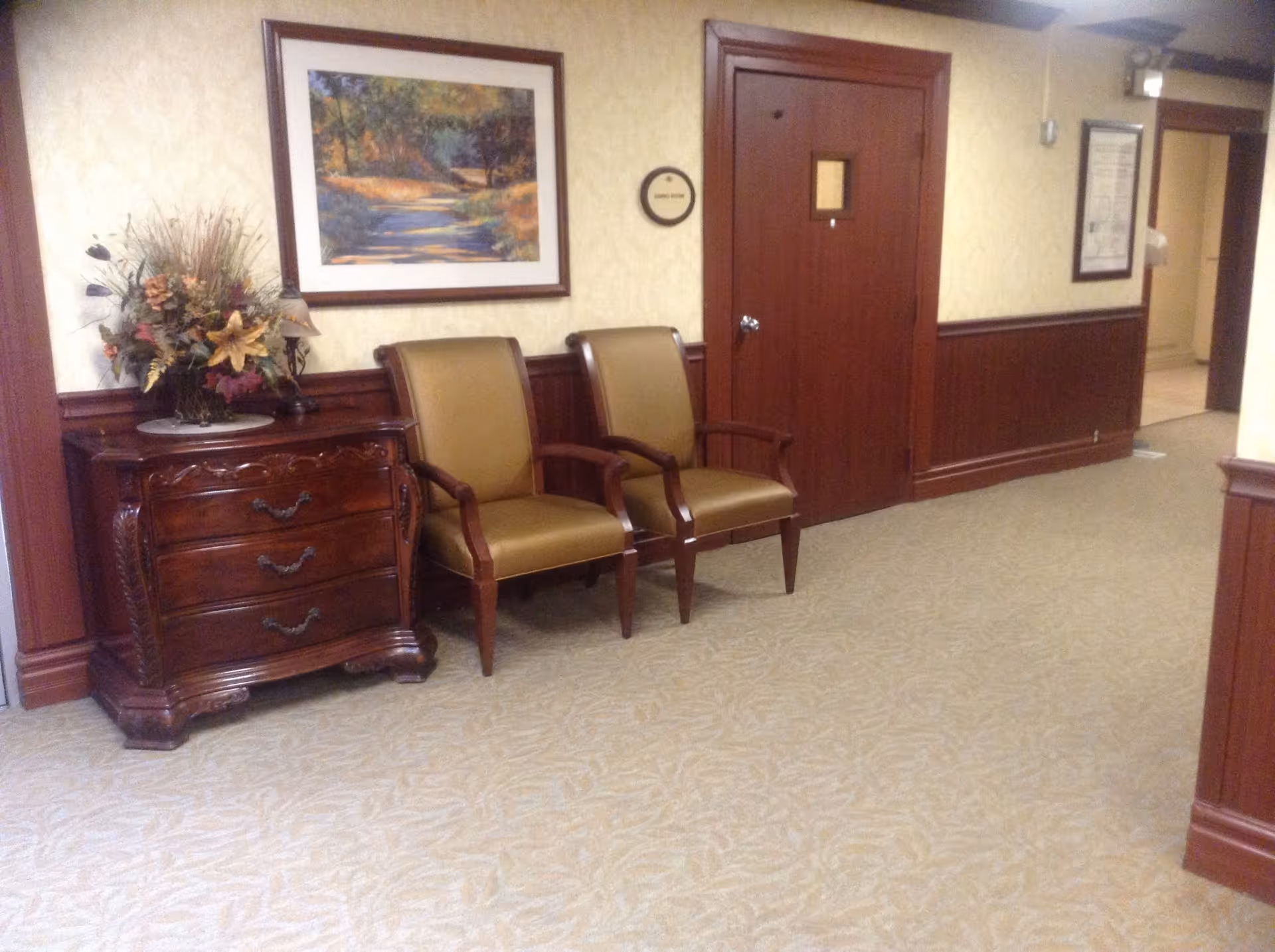 A hallway in The Claremont Hacienda facility with beige patterned carpet and cream-colored walls with wooden wainscoting. Two brown cushioned chairs are placed next to a dark wooden chest of drawers with a floral arrangement on top. A framed landscape painting hangs above the chairs. There is a closed wooden door with a small window and a round sign next to it, and an open doorway leading to another area.