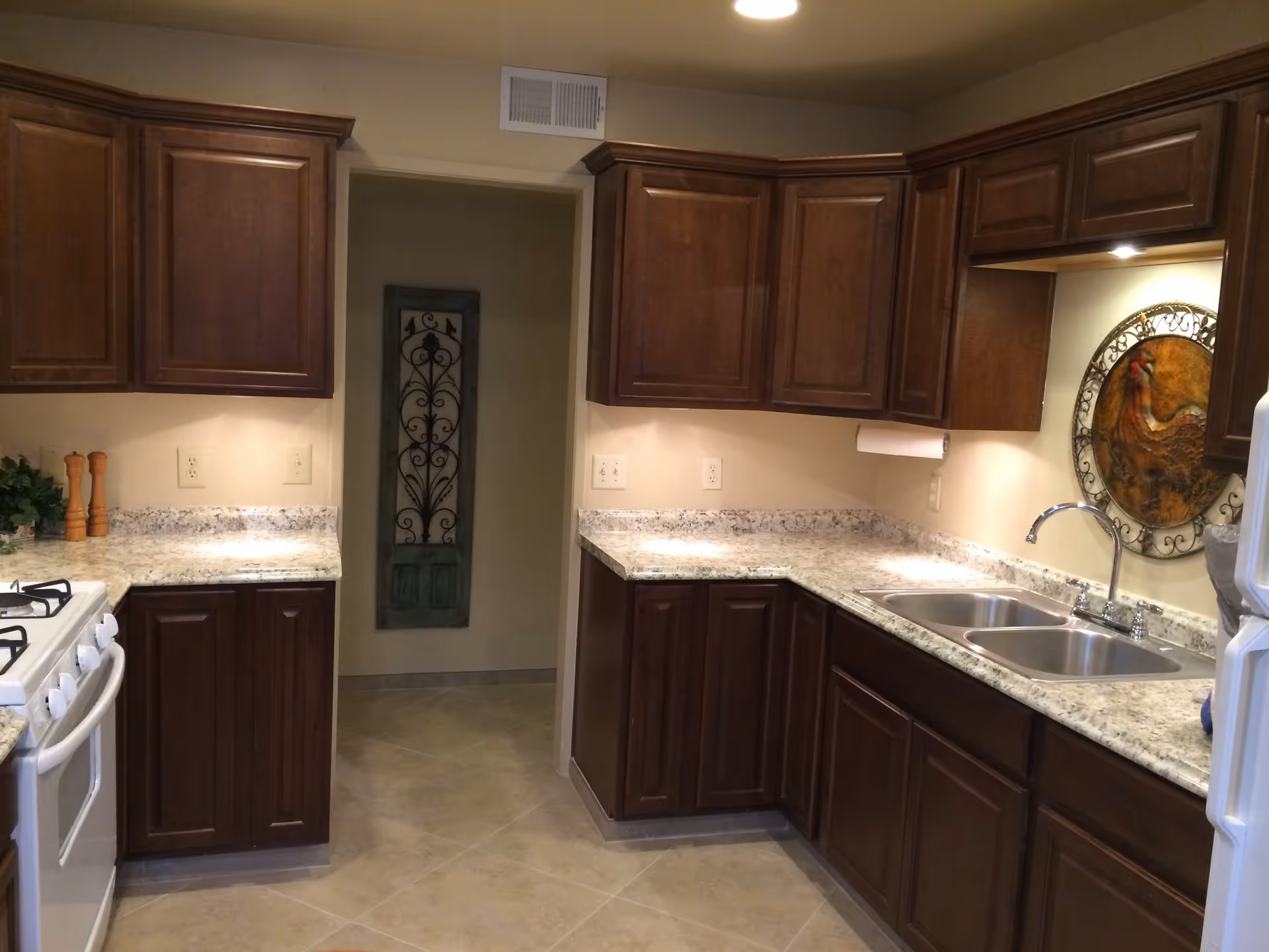 A kitchen with dark wooden cabinets, granite countertops, a double stainless steel sink, a white gas stove, and a decorative wall hanging featuring a rooster. The floor is tiled, and there is a doorway with a decorative metal panel on the wall inside the adjacent room.