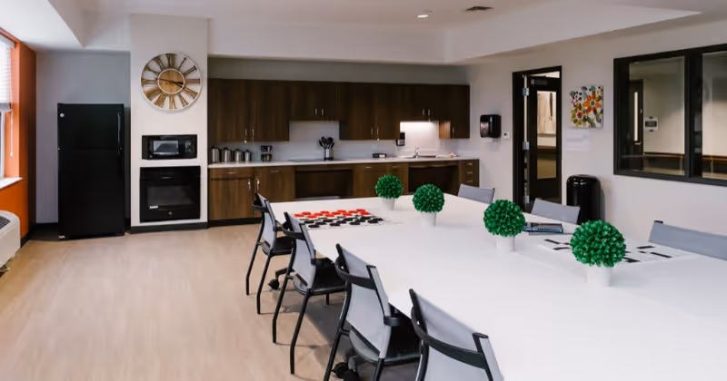 A modern communal kitchen and dining area with a long white table surrounded by chairs. The kitchen features dark wood cabinets, a black refrigerator, a microwave, and an oven. The table is decorated with small green potted plants and some documents. The room has light wood flooring and white walls with a large clock on one wall and a window letting in natural light.