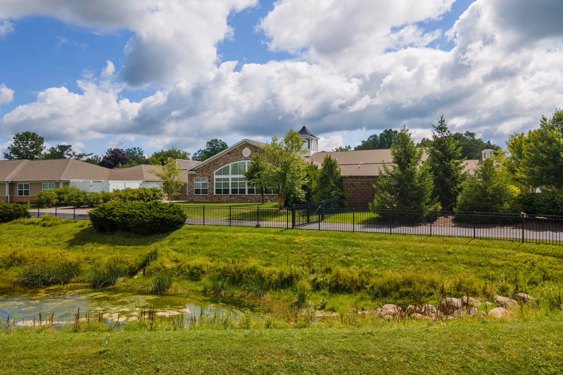 Exterior view of a senior living facility building with a large window, surrounded by green grass, bushes, trees, and a small pond under a partly cloudy sky.