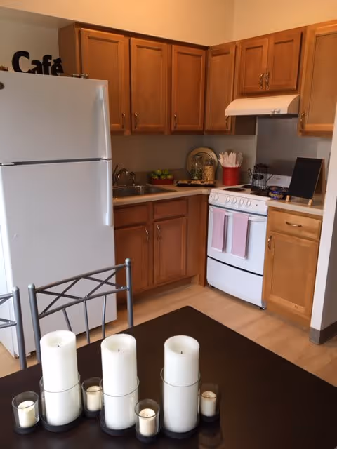 A small kitchen area with wooden cabinets, a white refrigerator, a white stove with two pink towels hanging on the handle, and a countertop with various items including a red container holding utensils. In the foreground, there is a dark table with several white candles of different sizes arranged on it.