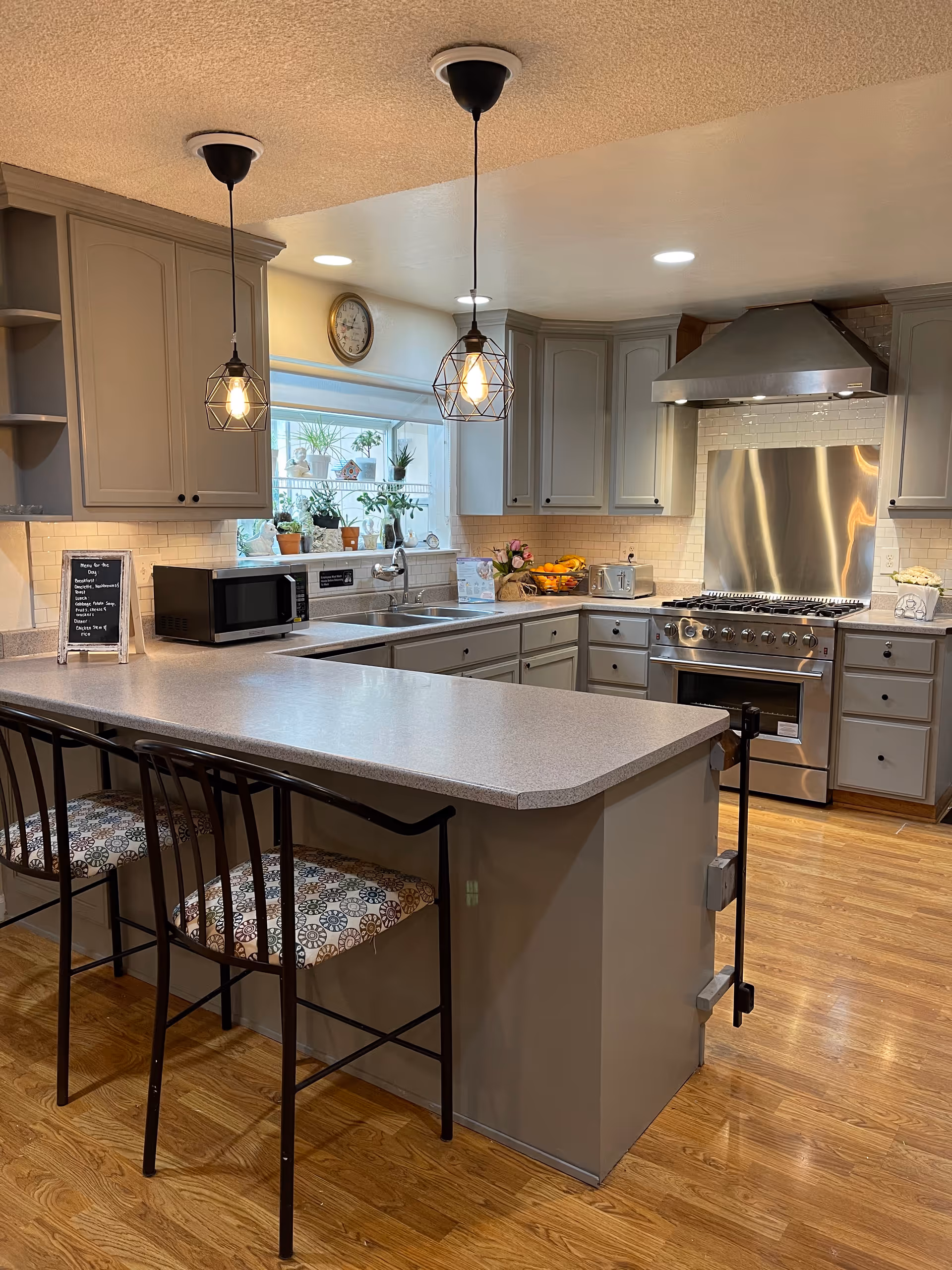 A modern kitchen with gray cabinets, a large countertop island with two patterned cushioned bar stools, stainless steel stove and range hood, a microwave, and a window with several potted plants on the sill. Two pendant lights hang from the ceiling above the island, and a clock is mounted on the wall above the window.