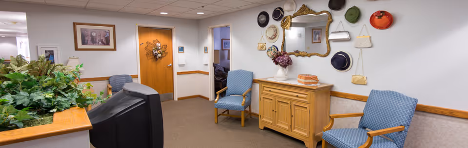 A cozy interior common area with two blue cushioned chairs, a wooden cabinet with decorative items on top, and a wall decorated with various hats and purses around a large ornate mirror. There is a TV in the foreground and a wooden door with a floral wreath in the background.