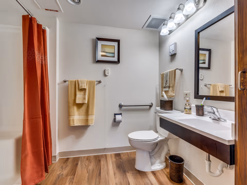 A clean and well-lit bathroom with a white toilet, a white sink with a dark wood vanity, a large mirror above the sink, and a red shower curtain. There are beige towels hanging on towel racks, a small framed picture on the wall, a tissue box, a soap dispenser, and a toothbrush holder on the sink. The floor has wood-like vinyl flooring.
