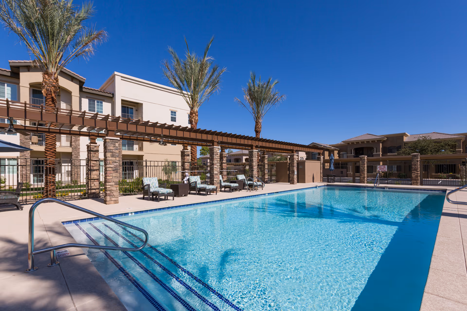 Outdoor swimming pool at The Enclave at Chandler Senior Living with clear blue water, surrounded by a concrete deck with lounge chairs and a pergola supported by stone columns. Palm trees and multi-story residential buildings are visible in the background under a clear blue sky.